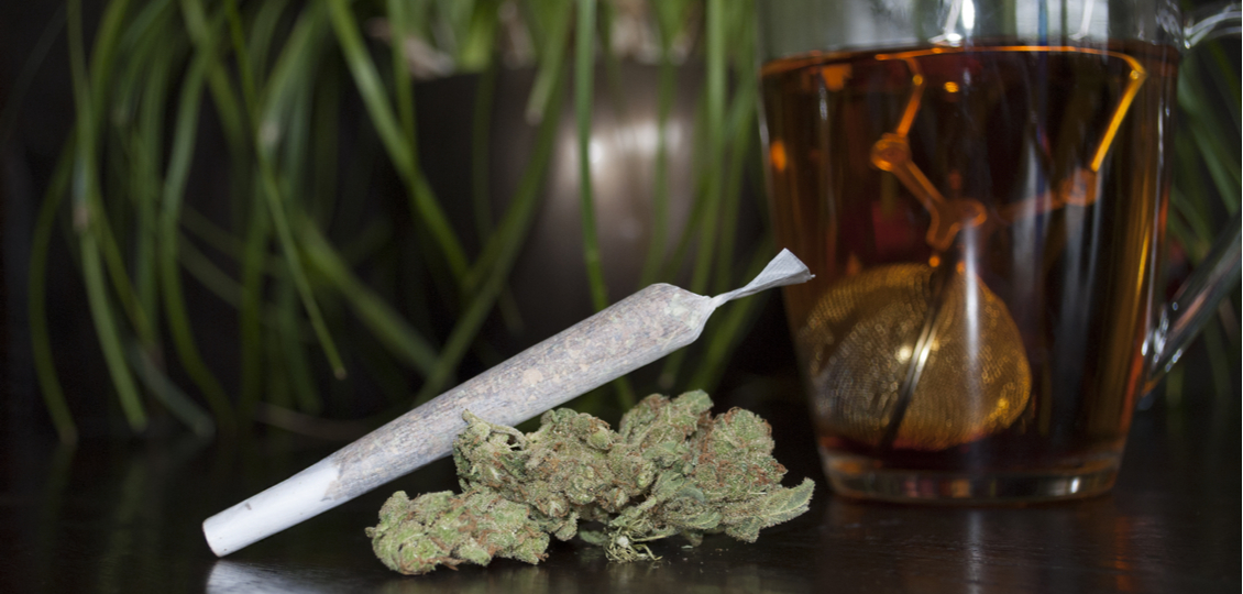 A joint leans on a dried cannabis flower with weed tea in the background